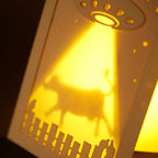 Laser cut cardboard art with a UFO and cow silhouette, illuminated from behind on a dark background
