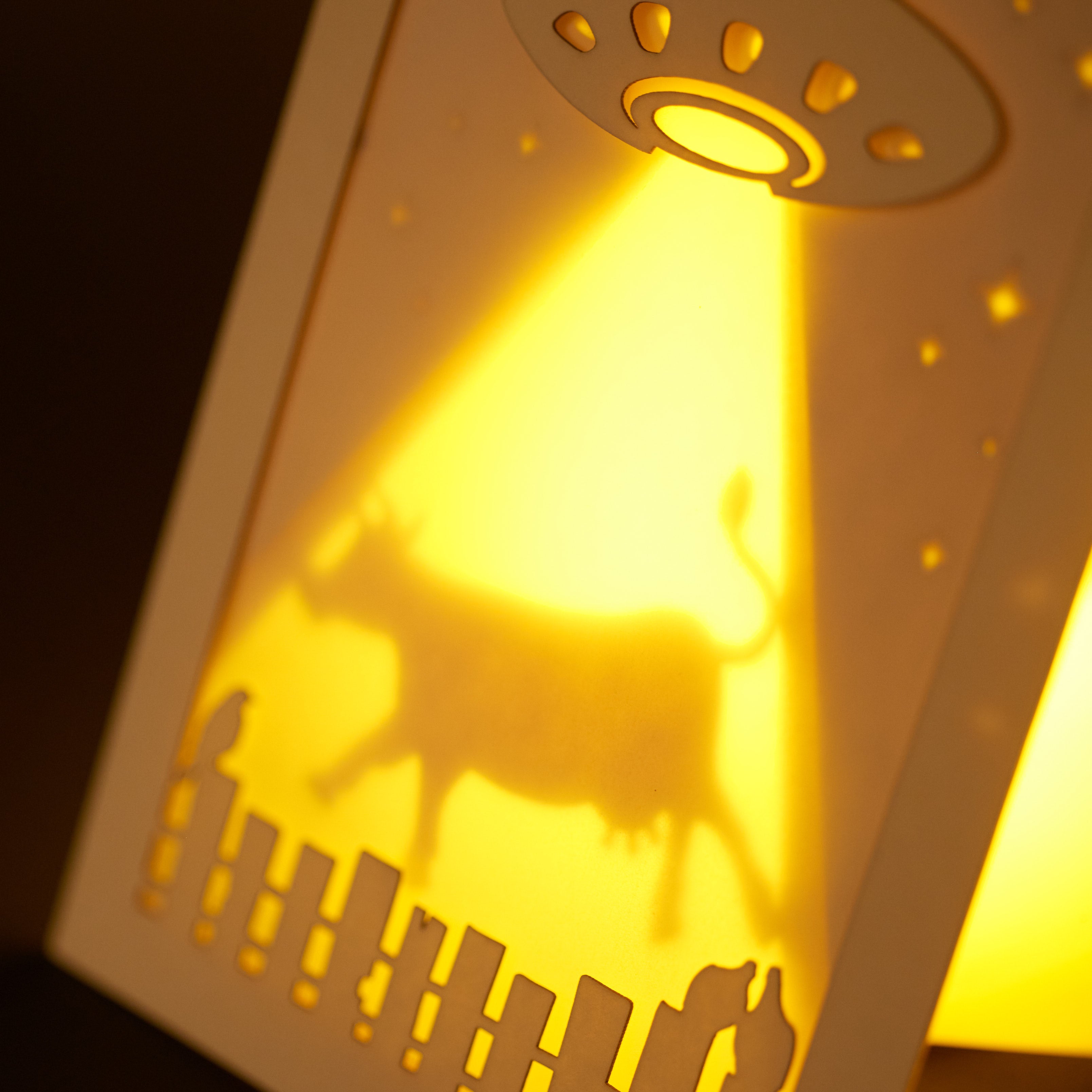 Laser cut cardboard art with a UFO and cow silhouette, illuminated from behind on a dark background