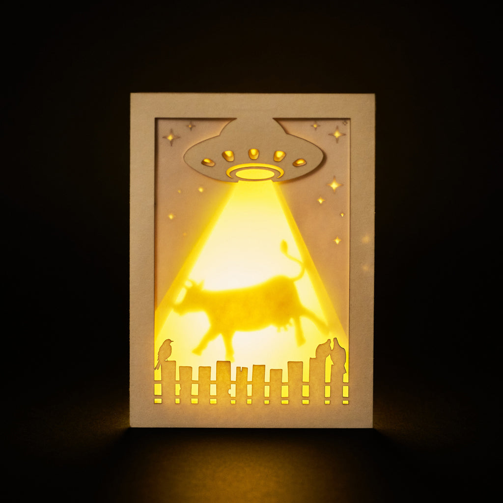 Laser cut cardboard art with a UFO and cow silhouette, illuminated from behind on a dark background