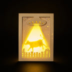 Laser cut cardboard art with a UFO and cow silhouette, illuminated from behind on a dark background
